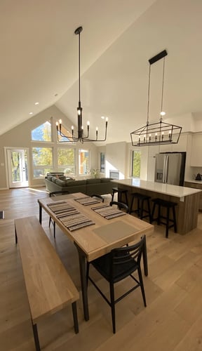 Open-plan kitchen, dining, and living room with vaulted ceiling, wood dining table, island counter, stainless steel appliances, and large windows overlooking