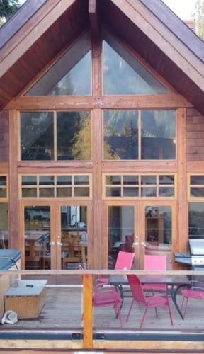Modern A-frame cabin with large glass doors, wooden deck with pink chairs and grill, surrounded by evergreen trees