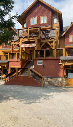 Red-brown wooden lodge building with large deck and wooden railings, adjacent shops at mountain village plaza