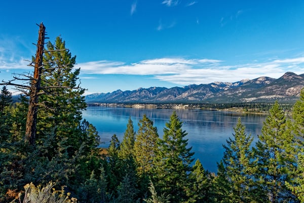 Modern mountain lodge with expansive windows overlooking forested valley, lake, and distant peaks at sunset
