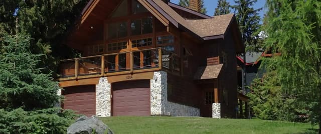 Modern A-frame cabin with dark wood siding, stone pillars, two-car garage, and wooden deck surrounded by evergreen trees and lawn.